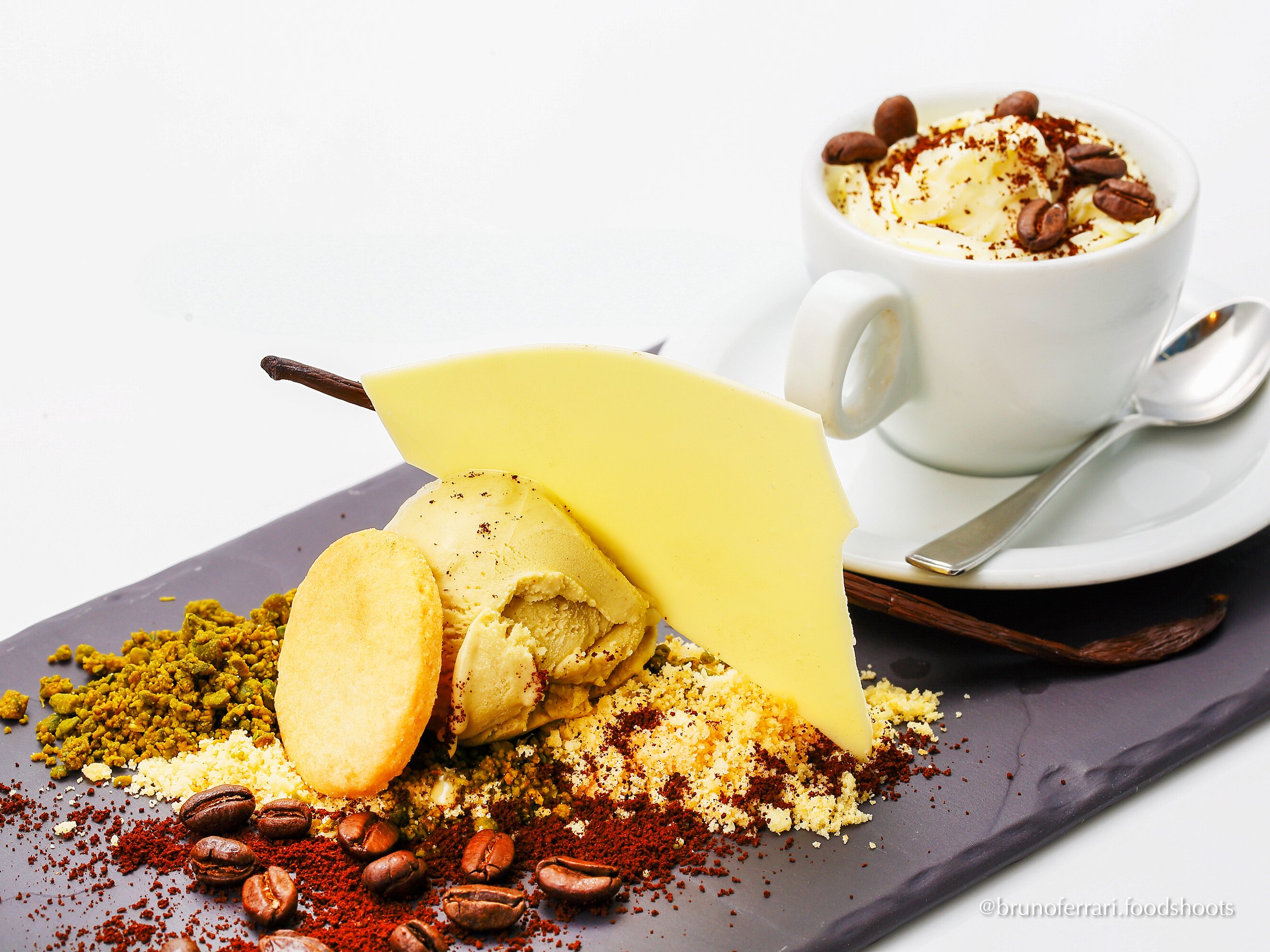 Dessert plate with ice cream, white chocolate, and cookie, next to a cup of whipped cream topped with coffee beans.