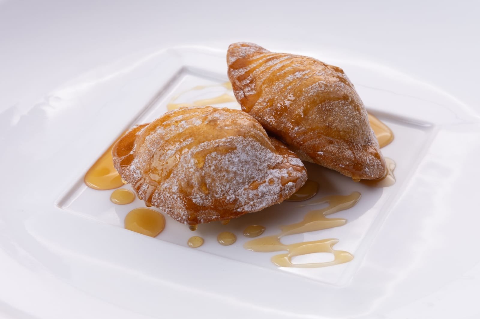 Two fried pastries dusted with powdered sugar and drizzled with caramel sauce on a white square plate. seadas sarda, special sardinia island dessert