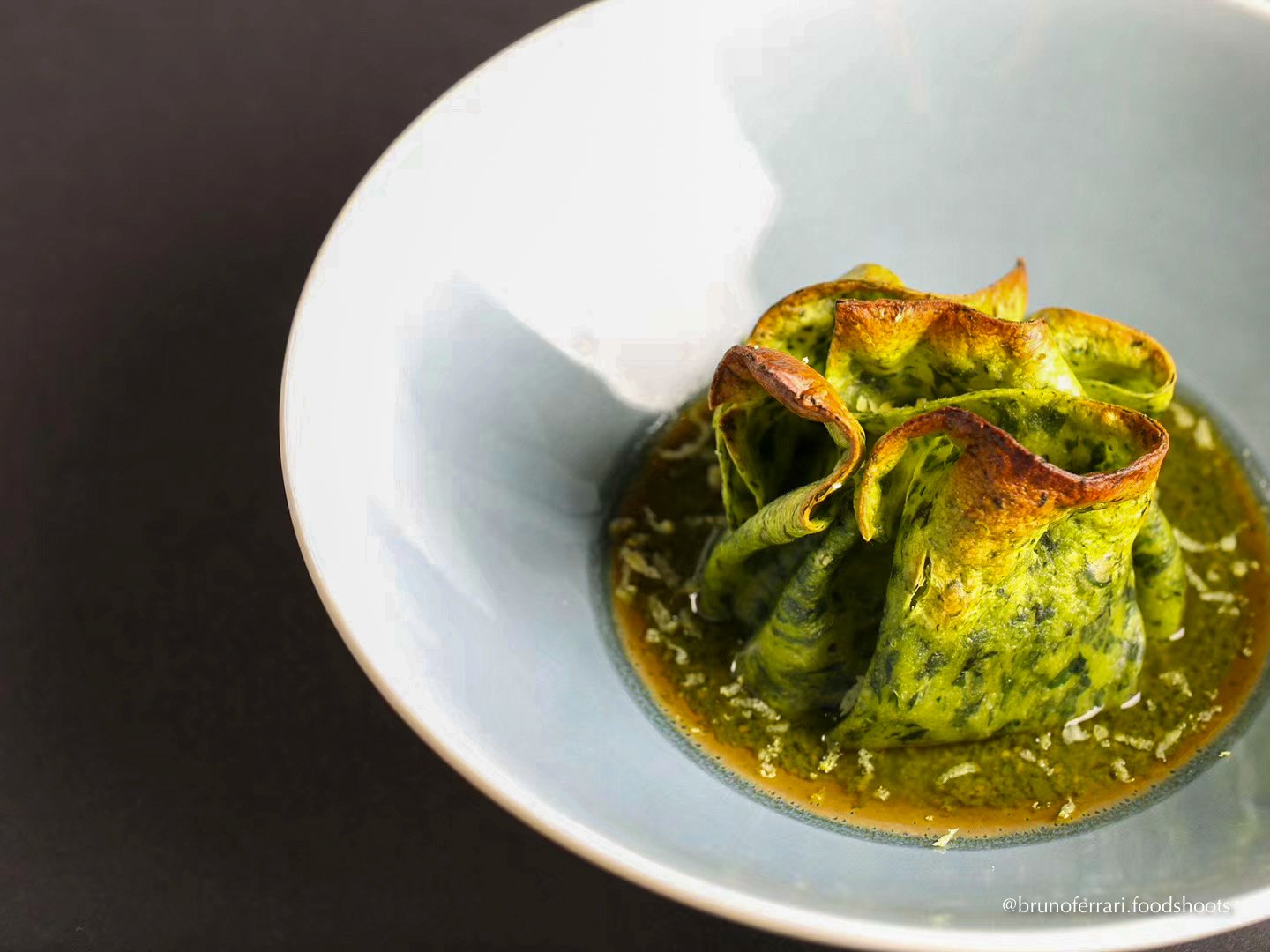 A white bowl containing a green vegetable, possibly zucchini or cucumber, cooked and rolled into a flower-like shape, in a green sauce or broth. lasagna green veghetarian basil pesto, potatoes, beans and bechamel sauce