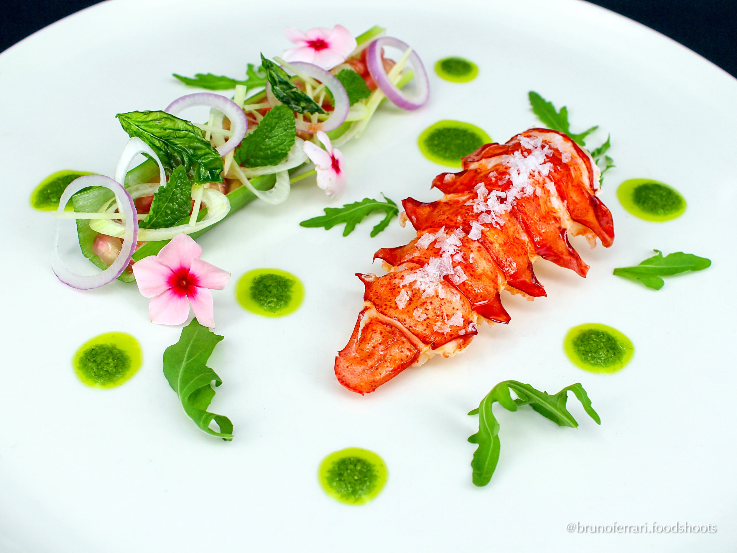 A plated dish featuring a lobster tail garnished with sea salt, served with a side of thinly sliced vegetables and herbs, decorated with pink flowers and green sauce dots. blue brittain lobster, fennel salad, basil pesto