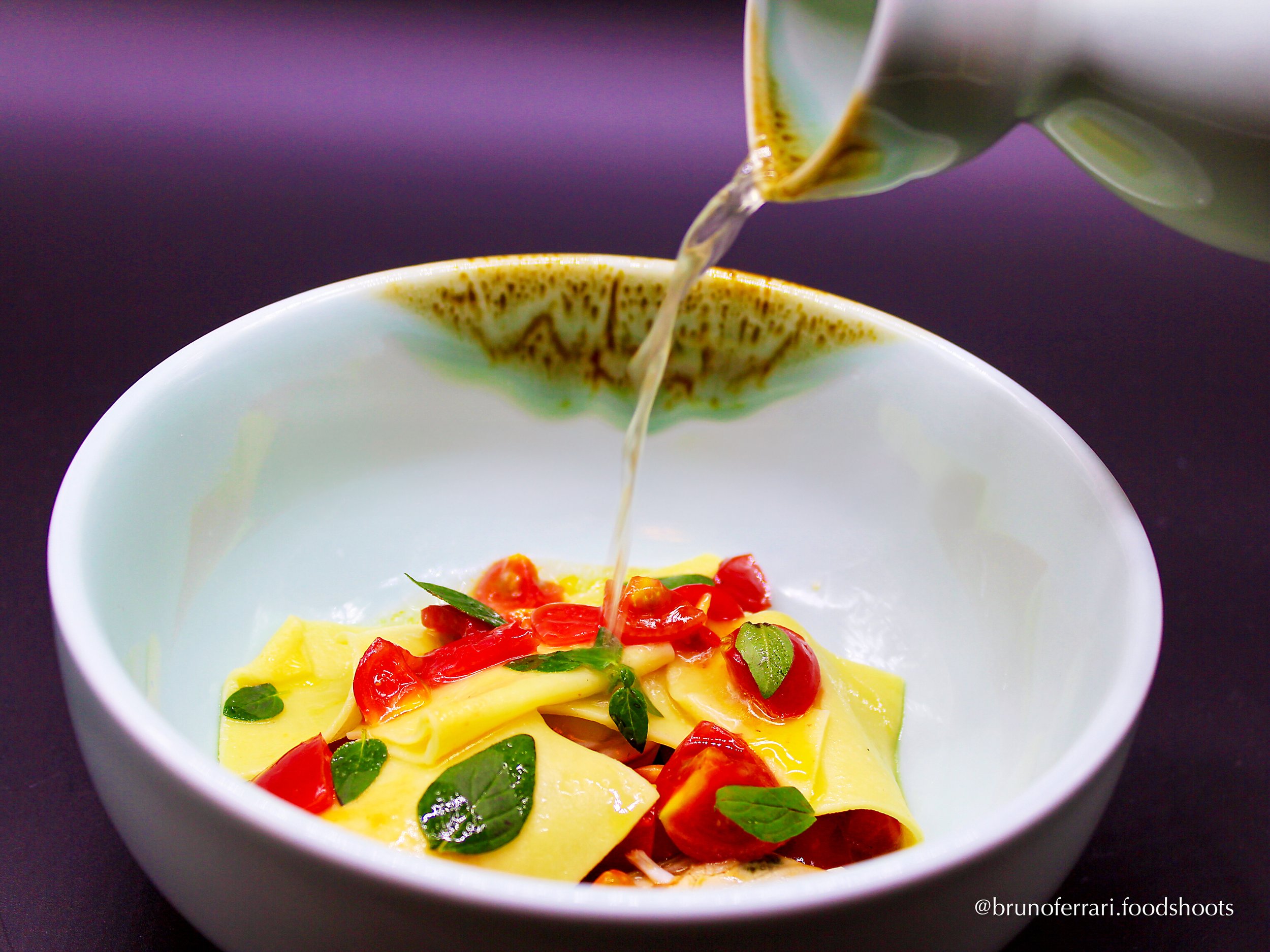 A bowl with fresh pasta, cherry tomatoes, and basil leaves, with oil being poured from a teapot.
