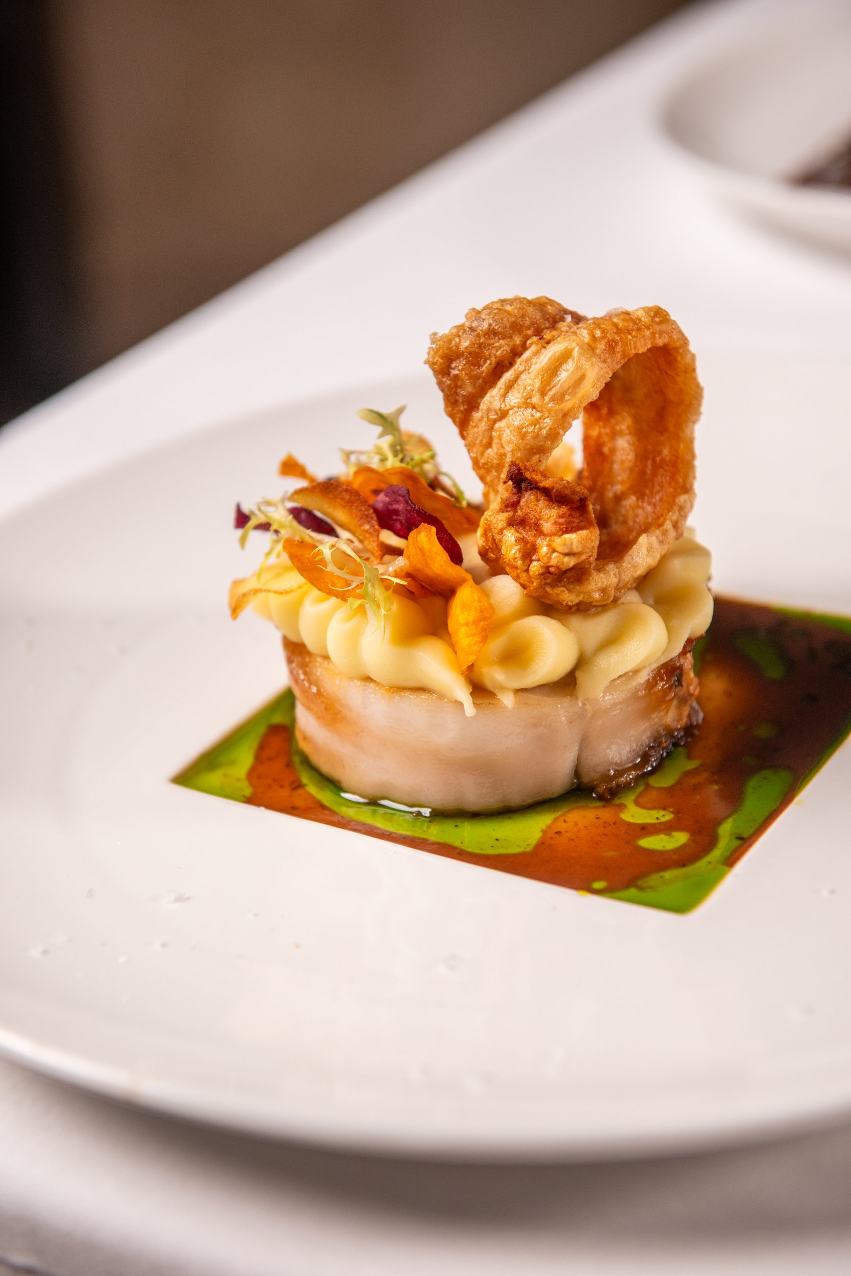A gourmet plated dish with a round piece of fish topped with mashed potatoes, crispy onion ring, edible flowers, and microgreens, all served with a drizzle of green oil and brown sauce on a white plate.