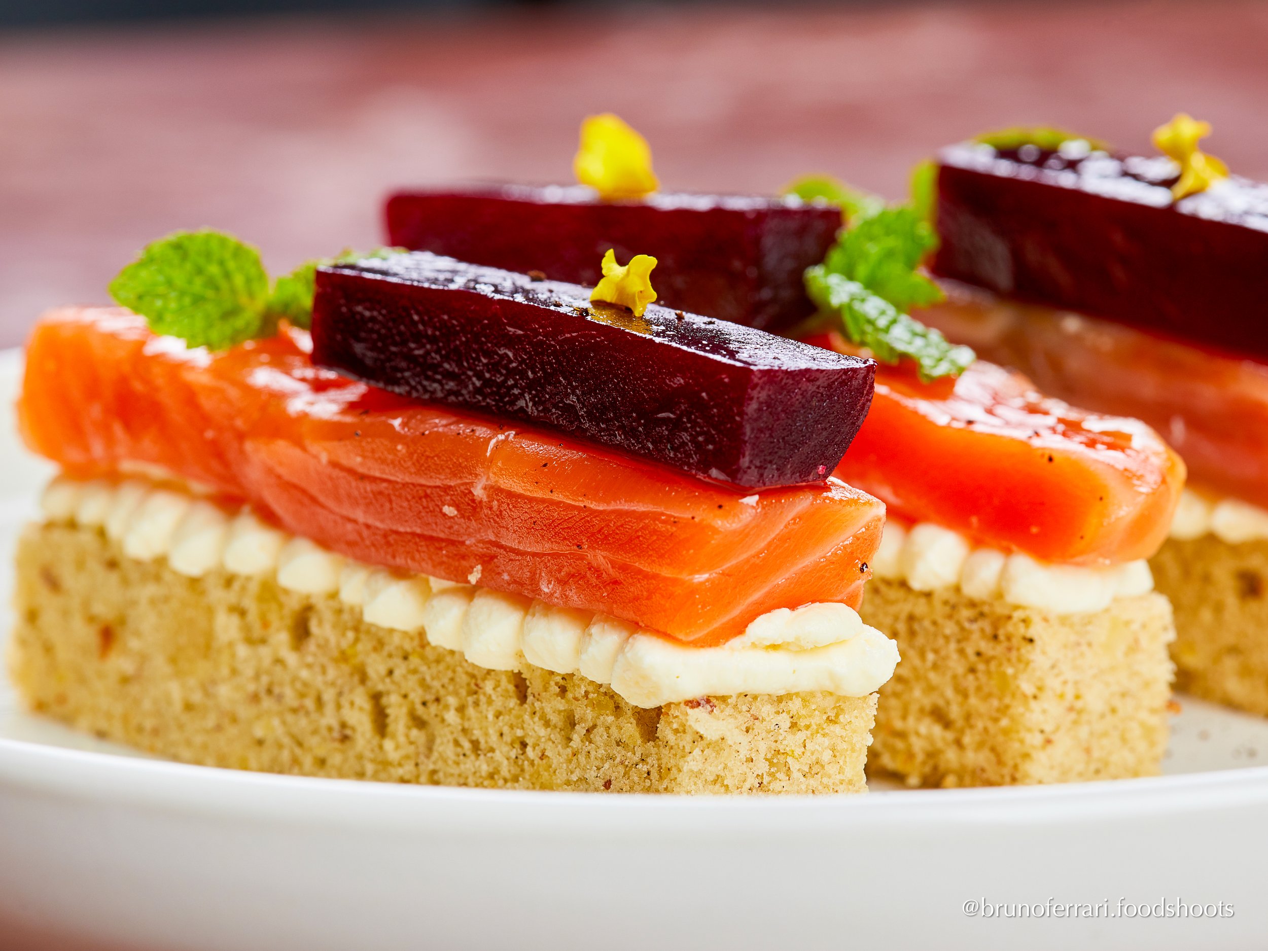 Appetizer with a rectangular piece of bread topped with cream cheese, smoked salmon, slices of beet, a mint leaf, and yellow flower garnish. gravlax salmon carpaccio, parrozzo bread, almonf, red beet
