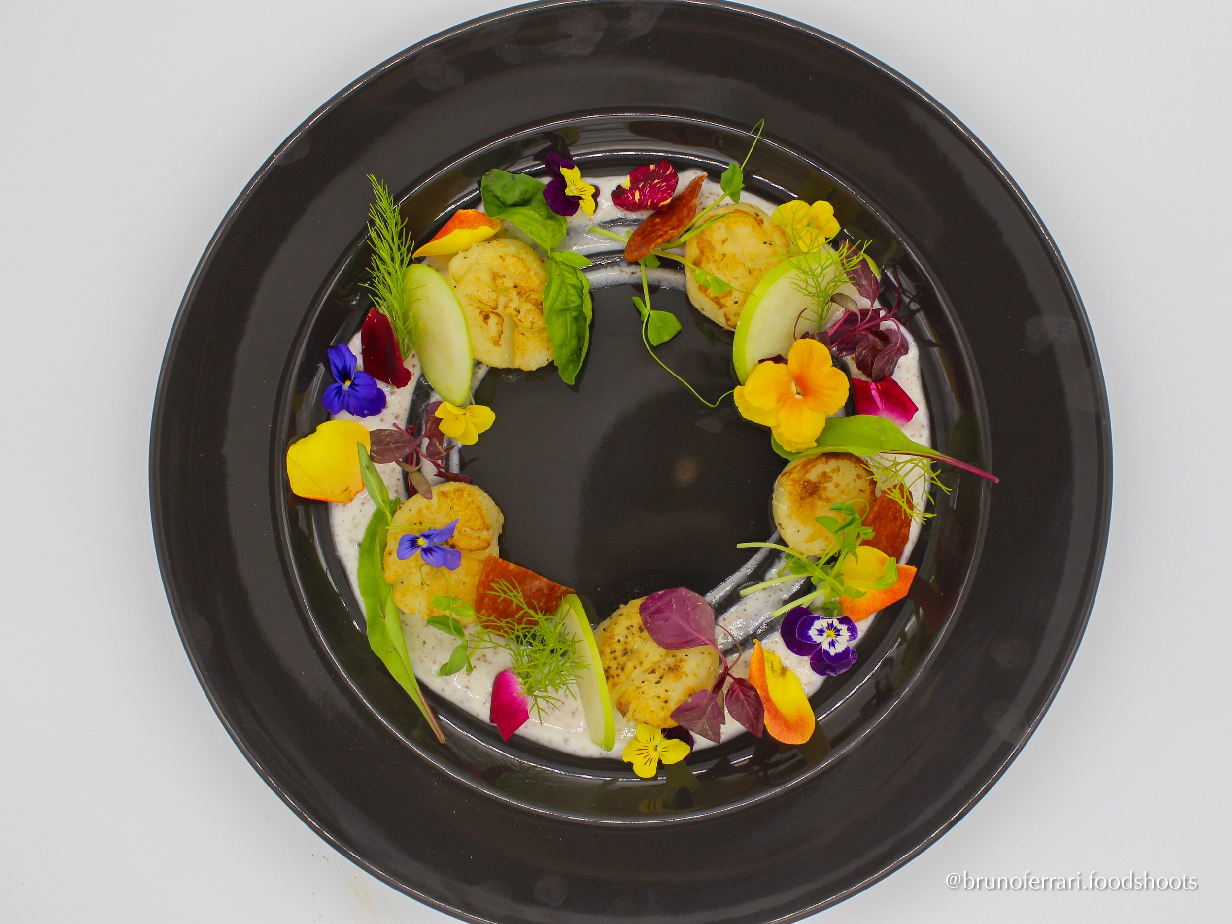 A black plate with a colorful arrangement of edible flowers, slices of green apple, seared scallops, and green herbs, artfully presented on a white background.