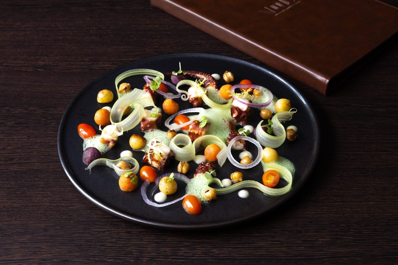 Gourmet dish with assorted small bites, onion rings, cherry tomatoes, microgreens, and foam, served on a black plate on a dark wooden table, with a menu or booklet in the background. octopus salad, sicilian style, celery, potatoes, black olives