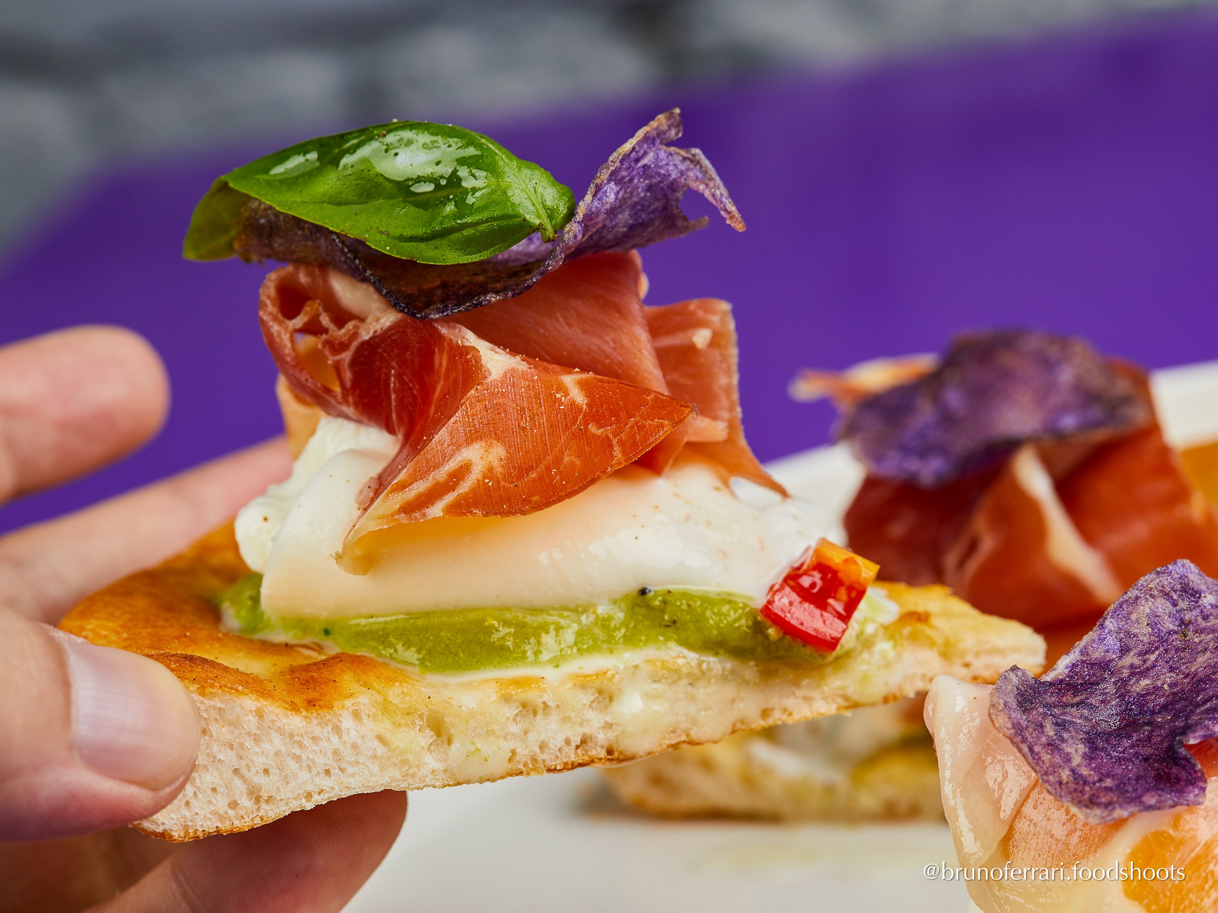 Close-up of an artisan appetizer featuring a thin pizza crust topped with cheese, sliced avocado, roasted red pepper, smoked salmon, a basil leaf, and purple potato chips, with a purple background. gourmet pizza, purple potatoes, spanish serrano ham