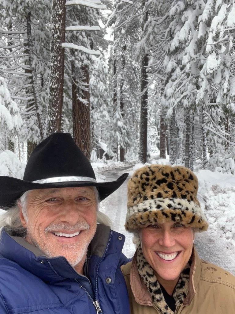 A smiling elderly man and woman taking a selfie in a snow-covered forest. The man is wearing a black cowboy hat and a blue jacket, and the woman is wearing a leopard print hat and tan jacket.