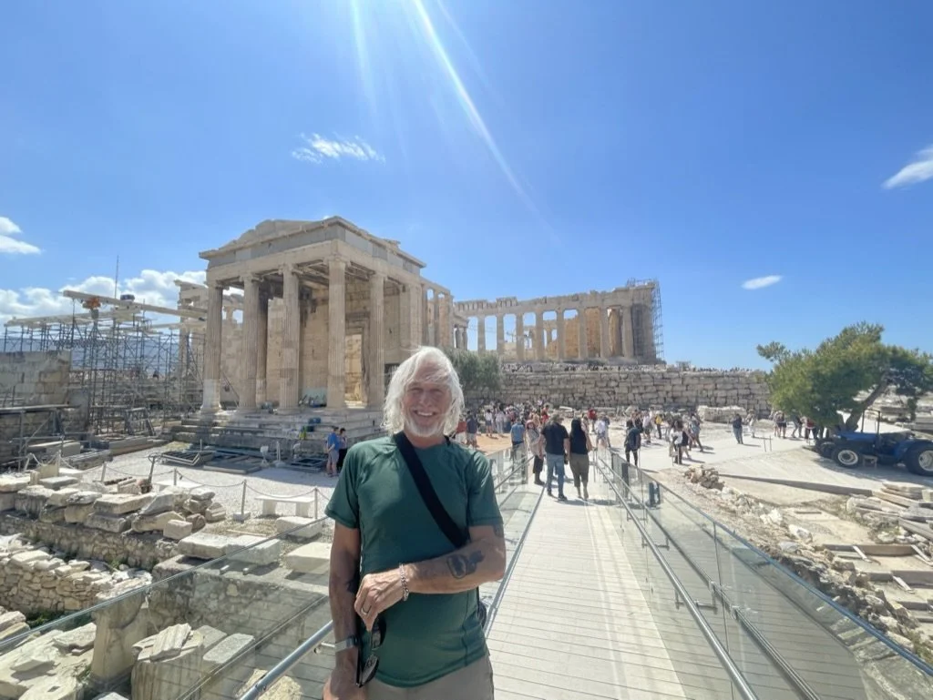 A smiling man standing on a walkway at the ancient Acropolis in Athens, Greece, with the Parthenon temple visible behind him and scaffolding on the side of the structure.