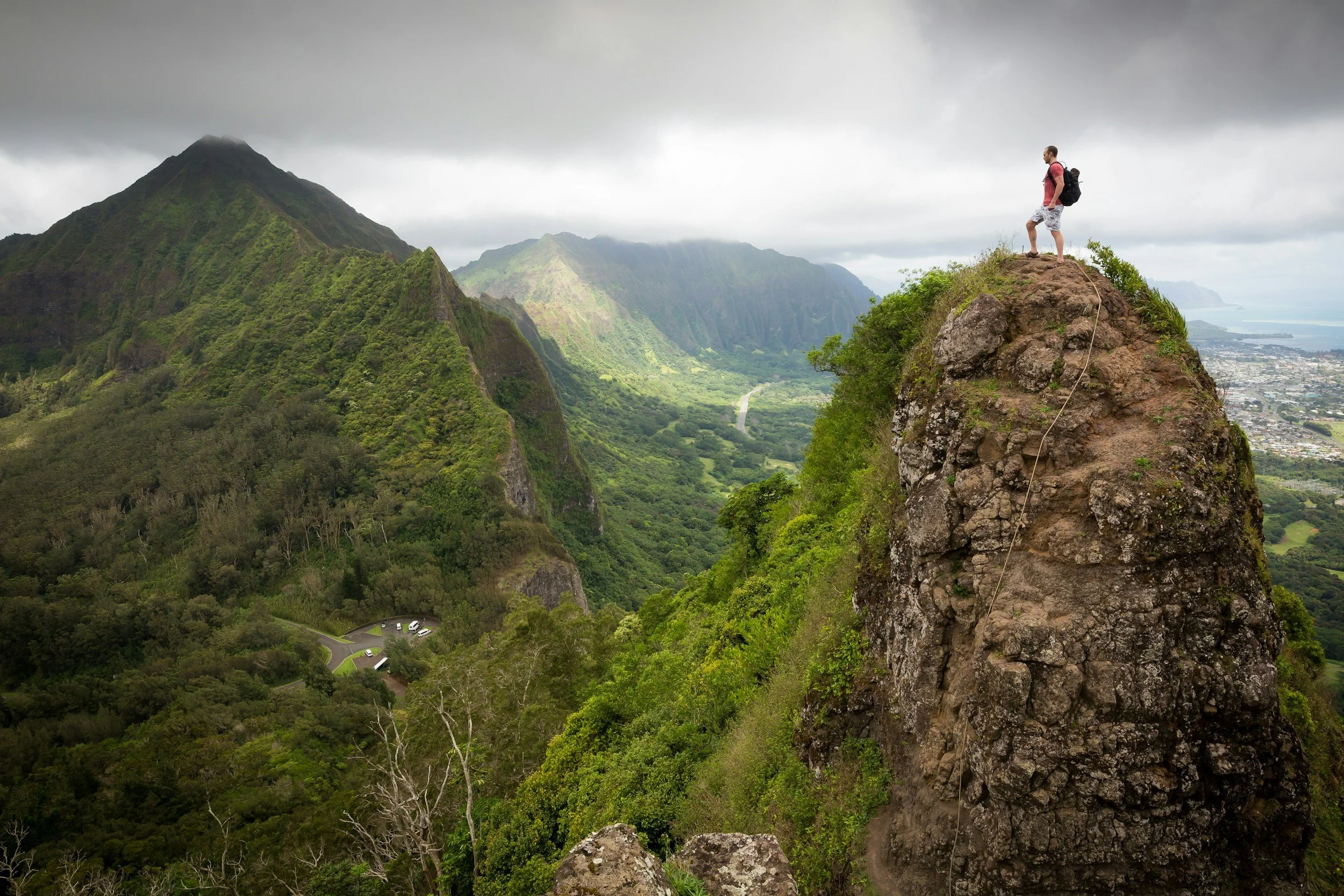 A man with a backpack standing on a large, rocky mountain peak overlooking a lush green valley and mountains with cloudy sky.