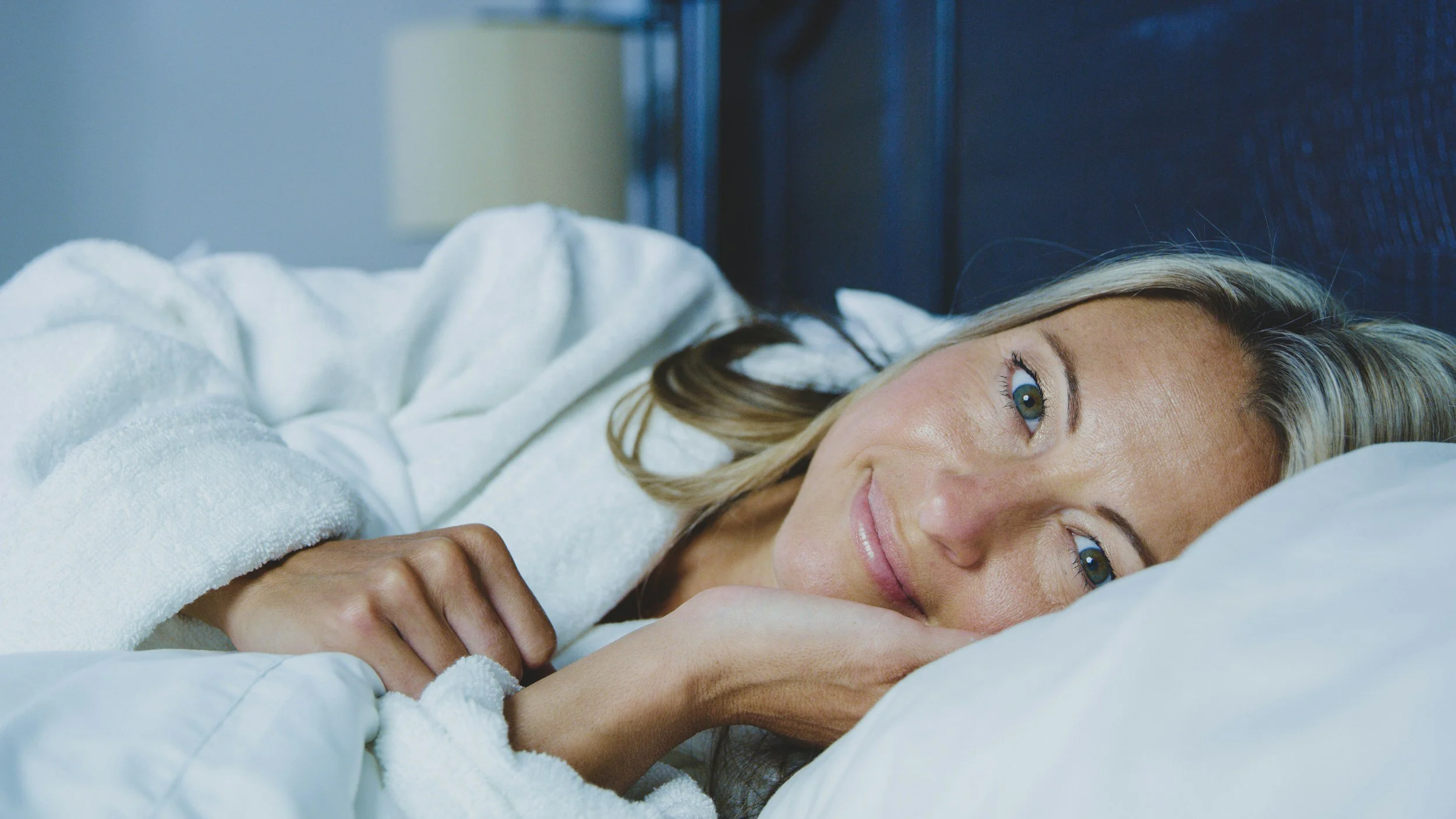 A woman with blonde hair lying on her side in bed, smiling, wearing a white bathrobe.