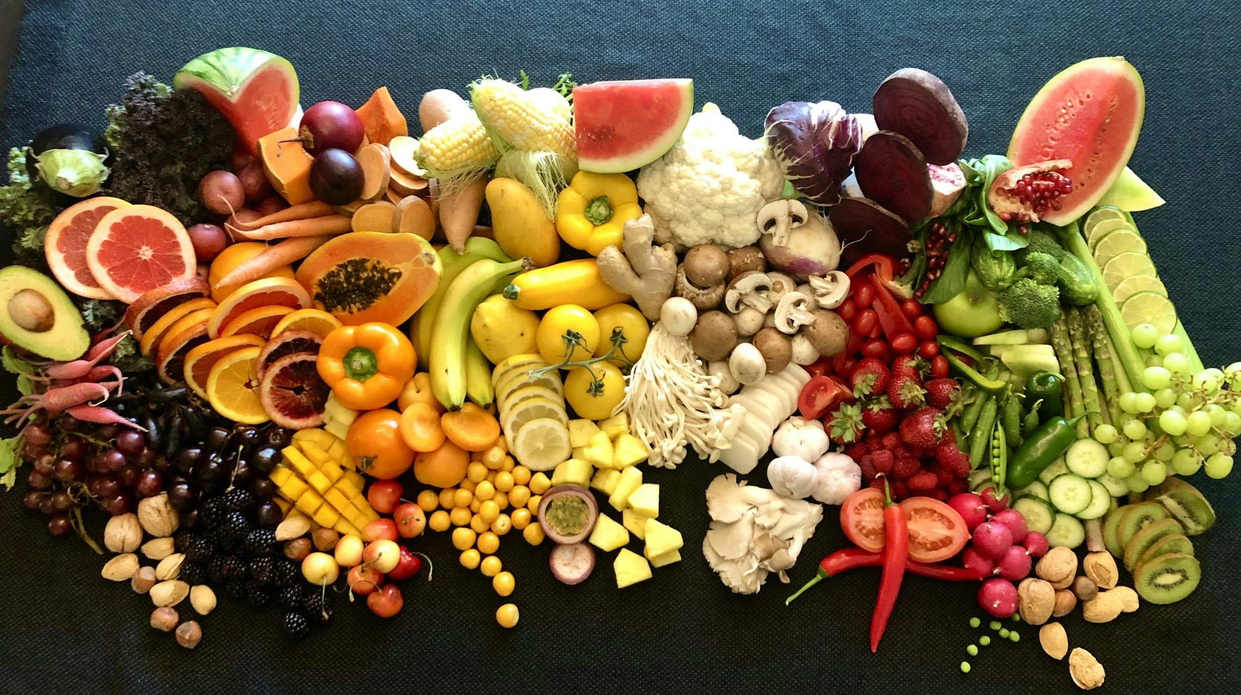 Assorted fresh fruits and vegetables arranged on a black surface, including watermelon, cauliflower, spinach, bell peppers, carrots, cherry tomatoes, strawberries, grapes, mushrooms, and citrus fruits.