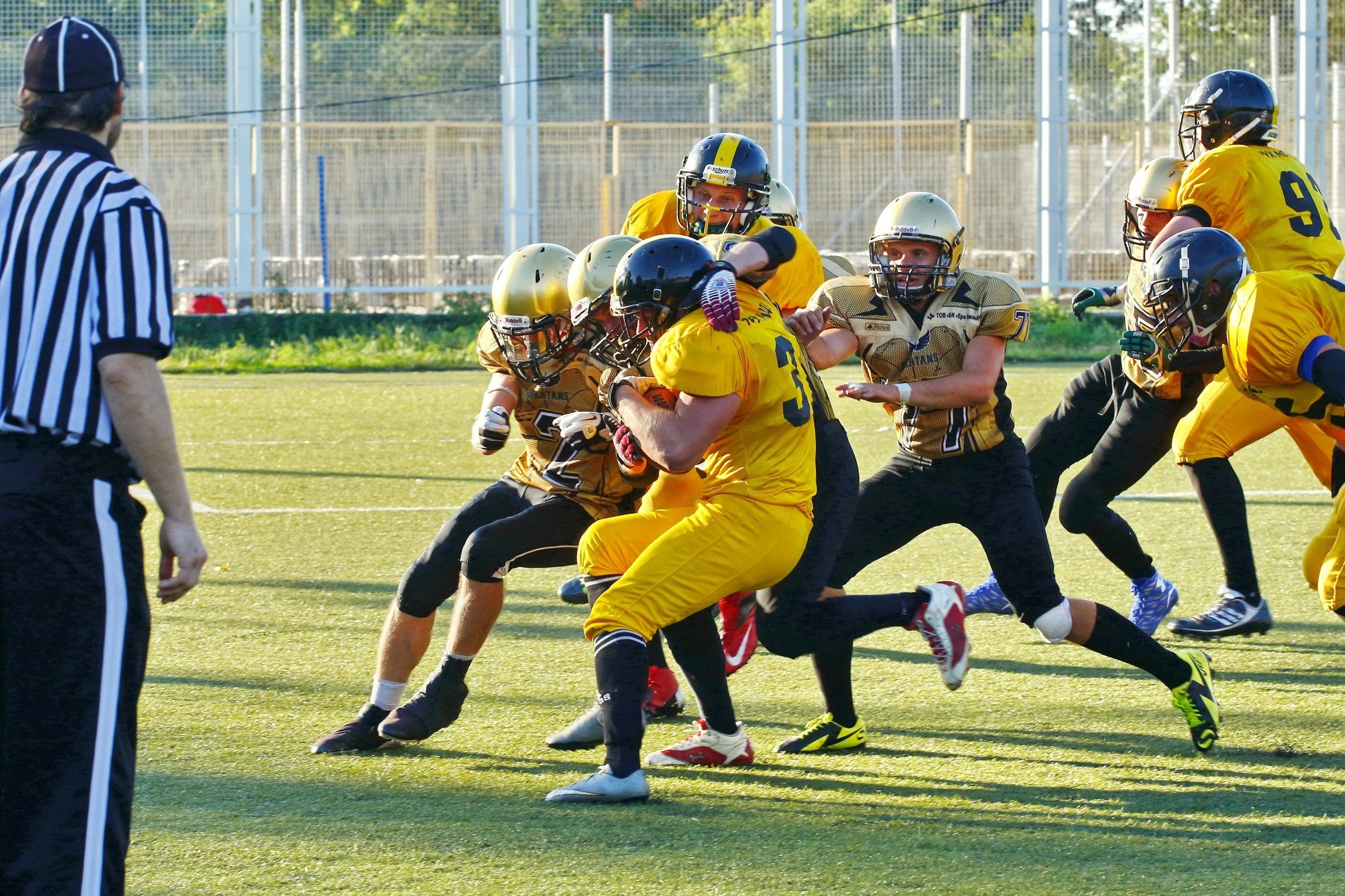 Youth football game with players in yellow and black uniforms, a referee in black and white stripes, and players tackling on the field.