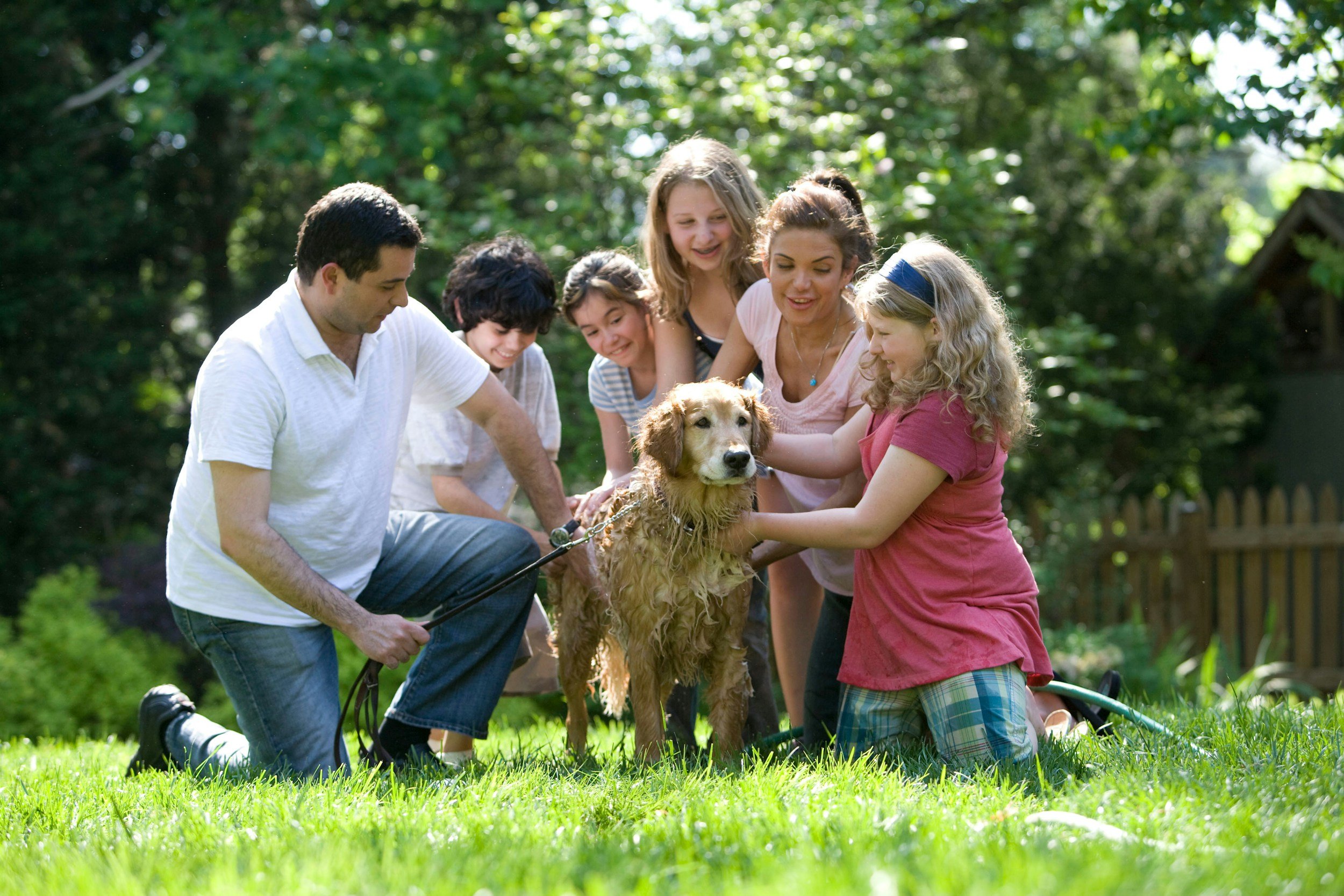 A group of people, including children and adults, gathered around a wet golden retriever dog outside in a grassy area with trees, smiling and petting the dog.