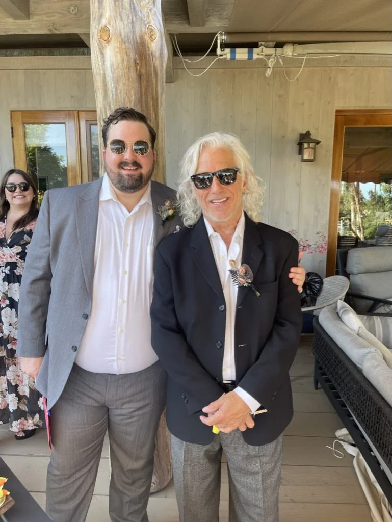 Three people standing together at a social gathering, outdoors in a rustic setting, dressed in semi-formal attire, wearing sunglasses, with a woman in the background smiling.