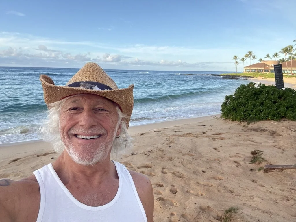 An older man with white hair and a beard taking a selfie on a sandy beach. He is wearing a straw cowboy hat, sunglasses, and a white sleeveless shirt. The ocean waves and blue sky are in the background, with some buildings and palm trees visible on the right side.