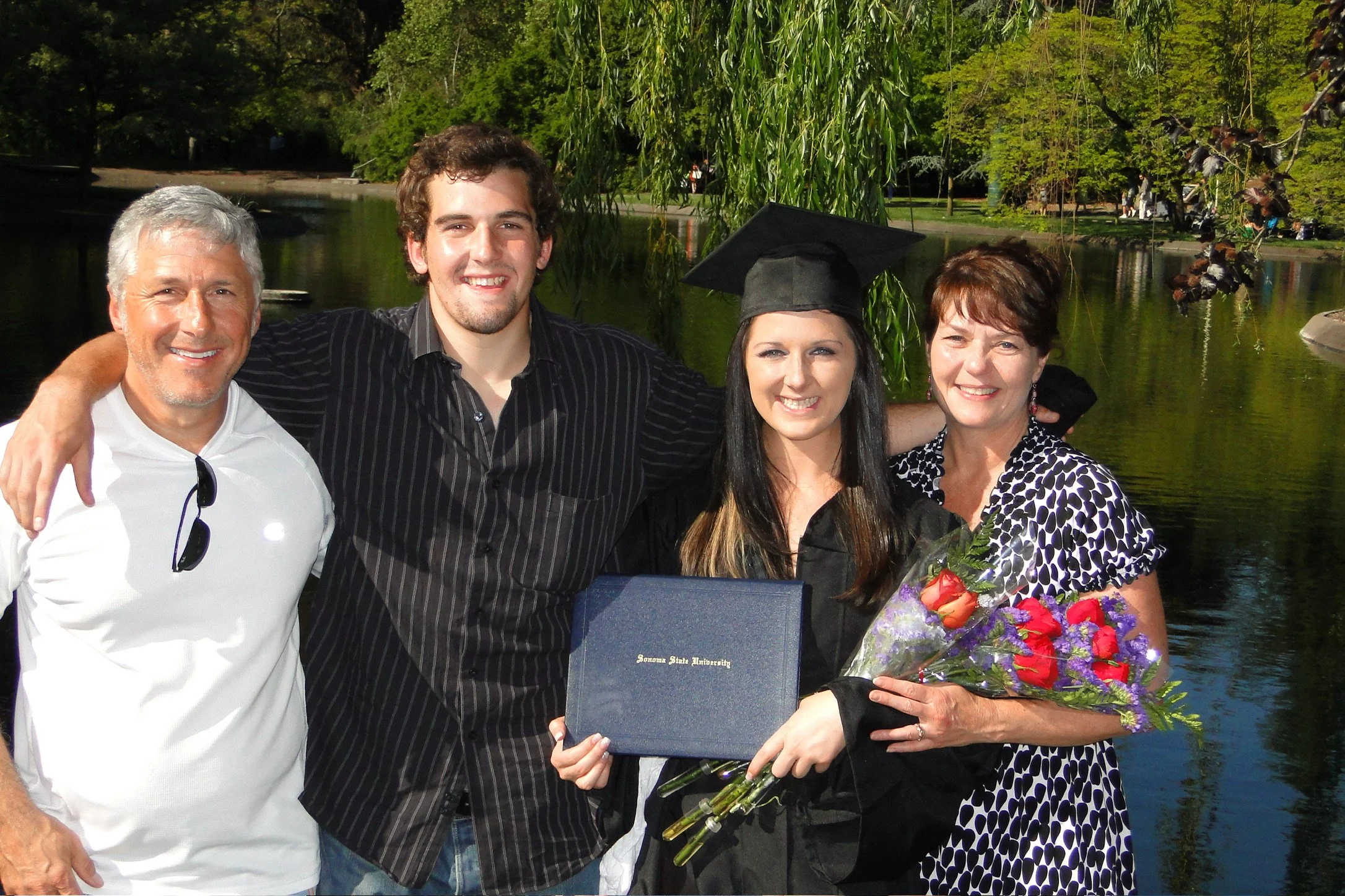 A woman in a graduation cap and gown holding a diploma and flowers, standing outdoors with three people, beside a lake and trees.