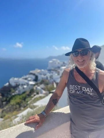 Smiling woman with blonde hair wearing sunglasses, a black hat, and a sleeveless gray shirt with the text 'Best Day of My Life', standing outdoors with a scenic coastal view of white buildings and the ocean in the background on a sunny day.