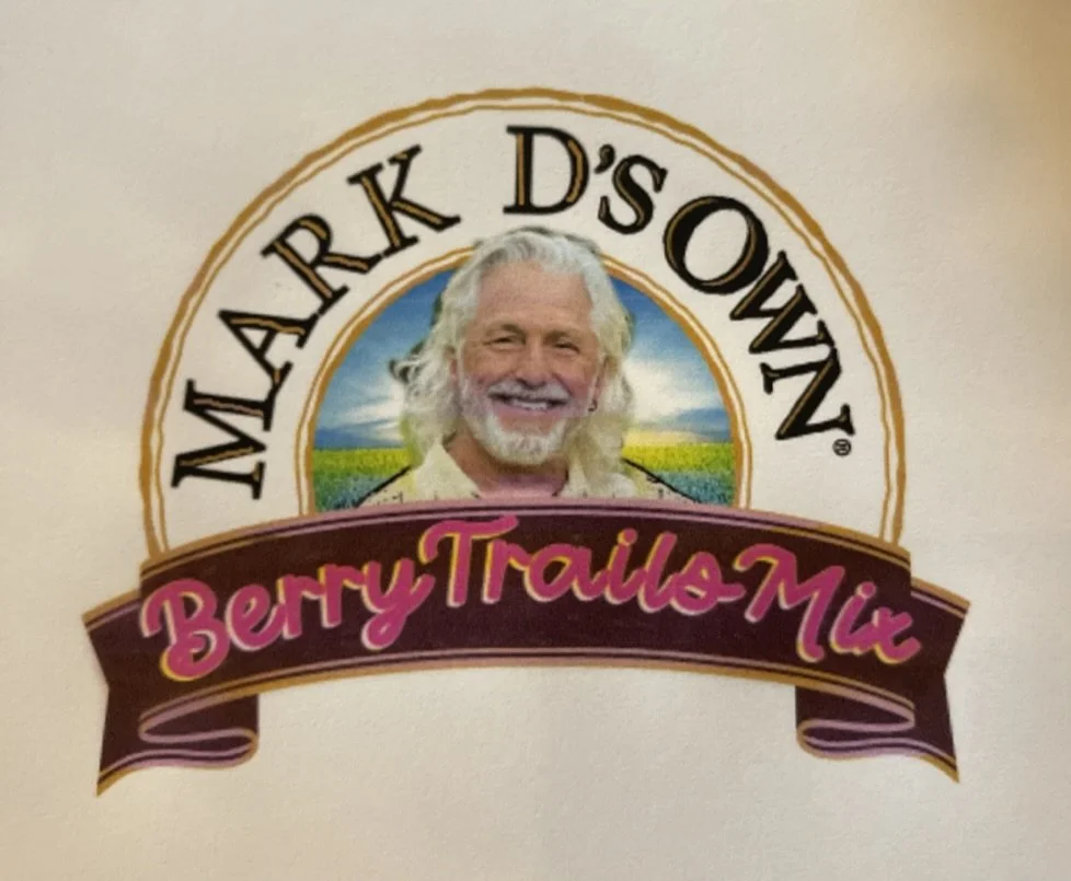 A sign for Mark D'sown Berry Trails Mix featuring a man with long white hair and a beard, smiling, with a landscape of fields and blue sky in the background.