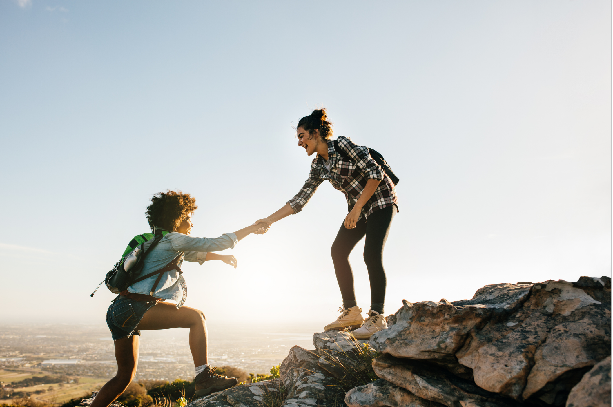 A woman helping a girl climb a rocky hill at sunset, both smiling and wearing backpacks.