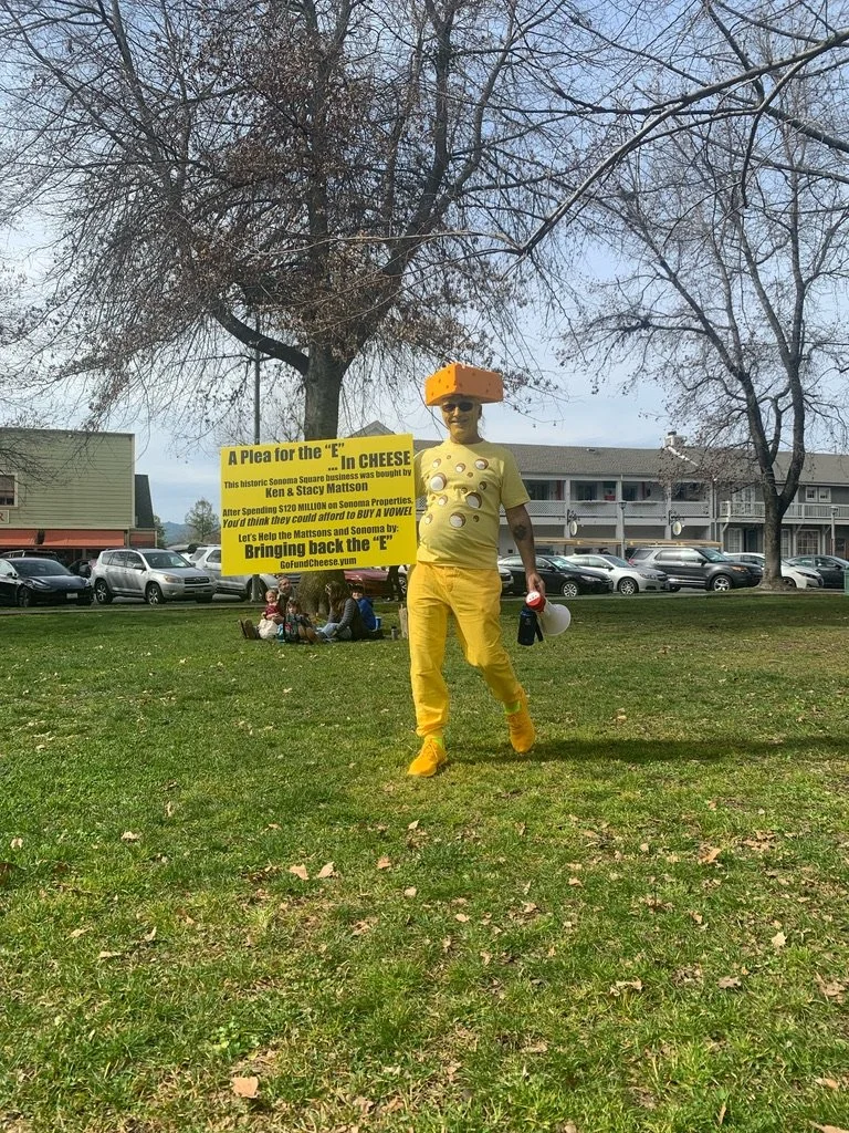 Person dressed as cheese with a cheese block on their head, holding a yellow sign about saving the 'E' in cheese, standing on grass in a park with leafless trees and buildings in the background.