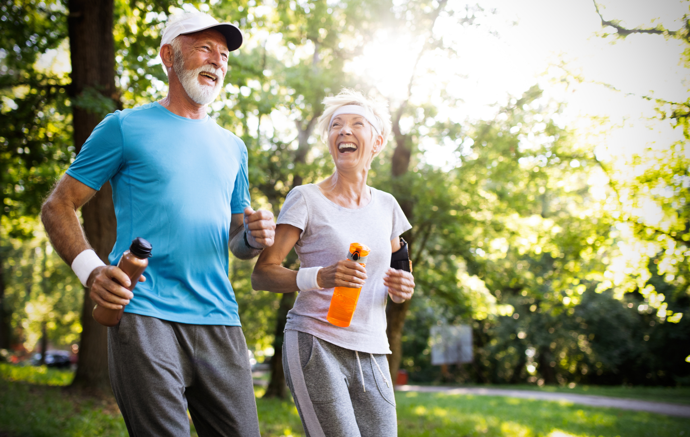 Older man and woman jogging together outdoors in a park, smiling and laughing, both holding water bottles, surrounded by green trees and sunlight.