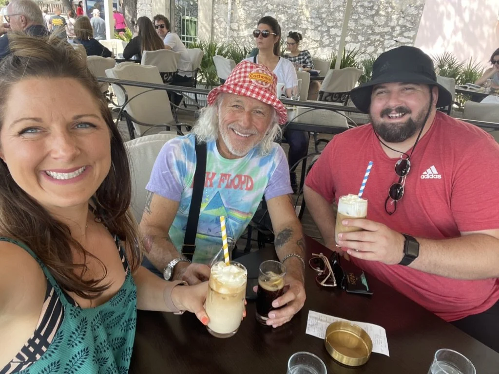 Three people smiling and sitting at a table in an outdoor cafe, each holding a drink. The woman on the left has long brown hair, the man in the middle has long gray hair and a red checkered hat, and the man on the right is wearing a black hat and sun