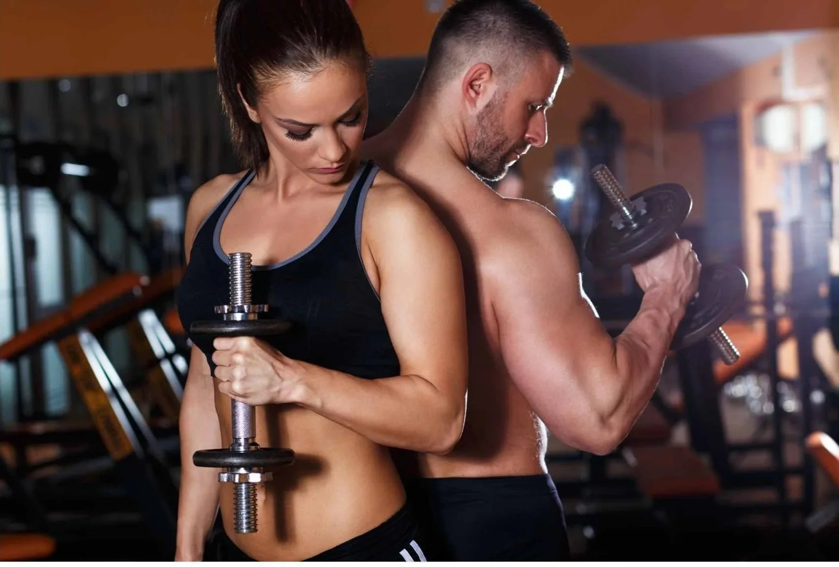 A woman and man in a gym lifting dumbbells, standing back to back in a focused pose.