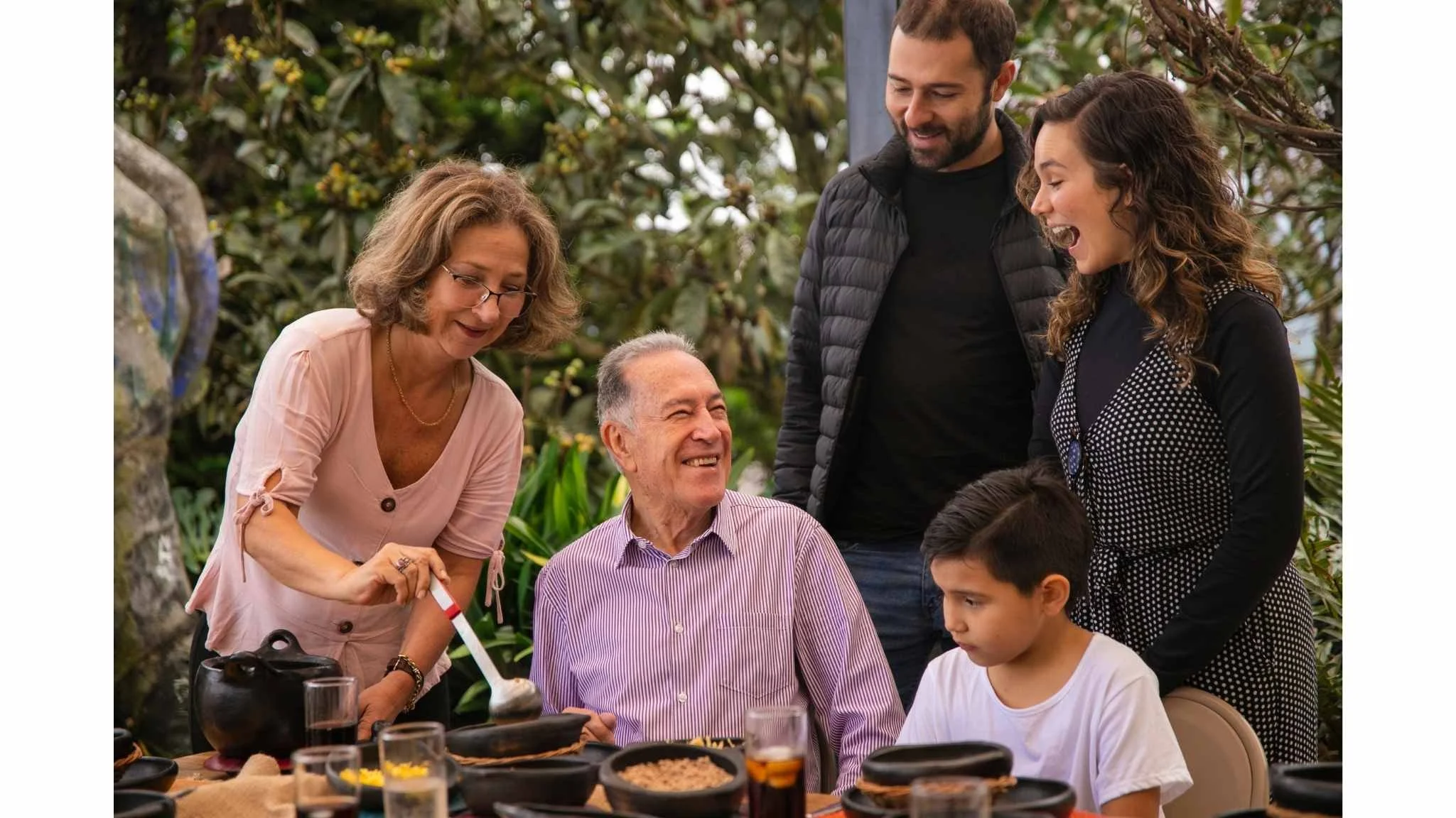 Family of six gathering outdoors at a table, smiling and enjoying a meal together, surrounded by lush greenery.