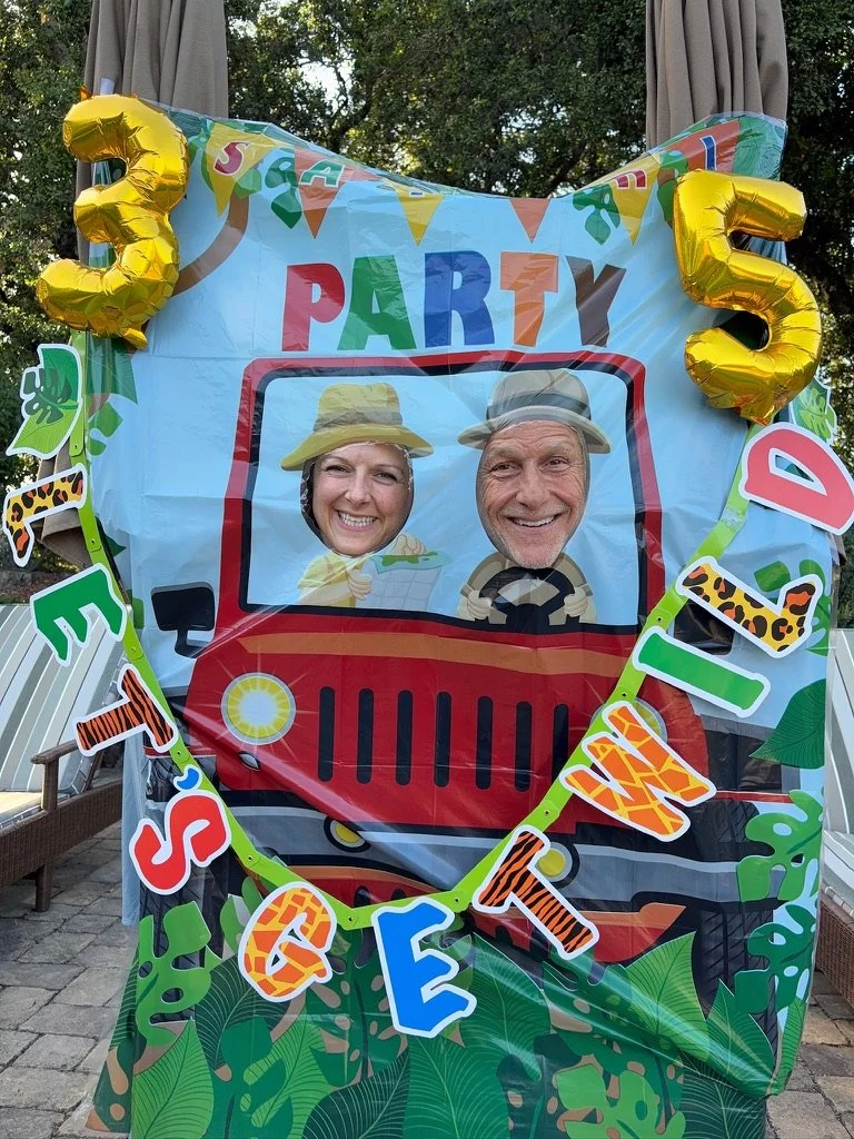 A photo booth picture at a jungle animal-themed birthday party. The scene features a colorful backdrop with a red Jeep and cartoon explorer faces with headgear, smiling faces of a man and a woman. The display includes gold foil balloons shaped as the