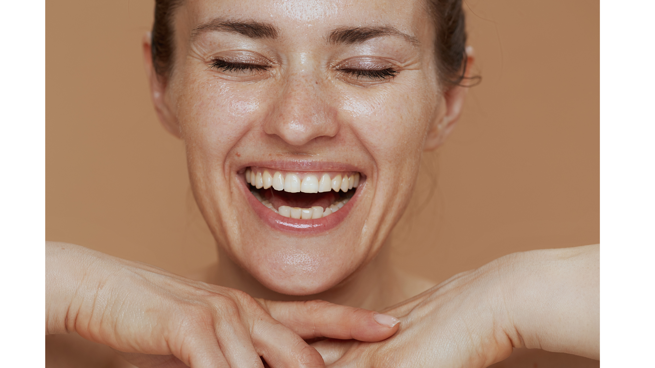 A smiling woman with closed eyes, showing her teeth, resting her chin on her hands against a beige background.