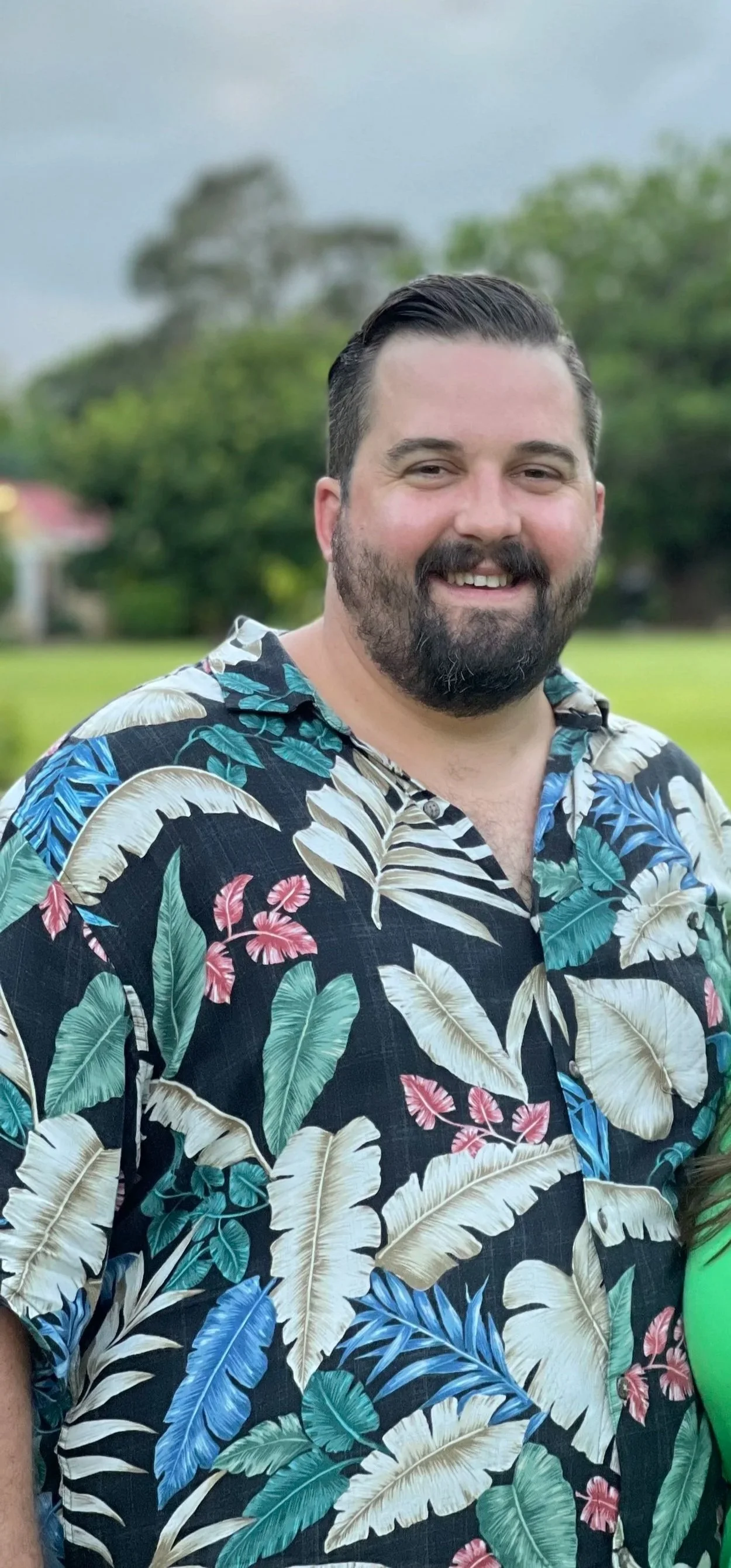 A smiling man with a beard and slicked-back hair wearing a colorful Hawaiian shirt standing outdoors with trees and a cloudy sky in the background.