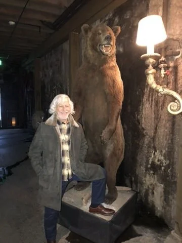 A woman with white hair and glasses standing next to a large bear statue in a rustic, wood-paneled room with a vintage wall-mounted lamp.