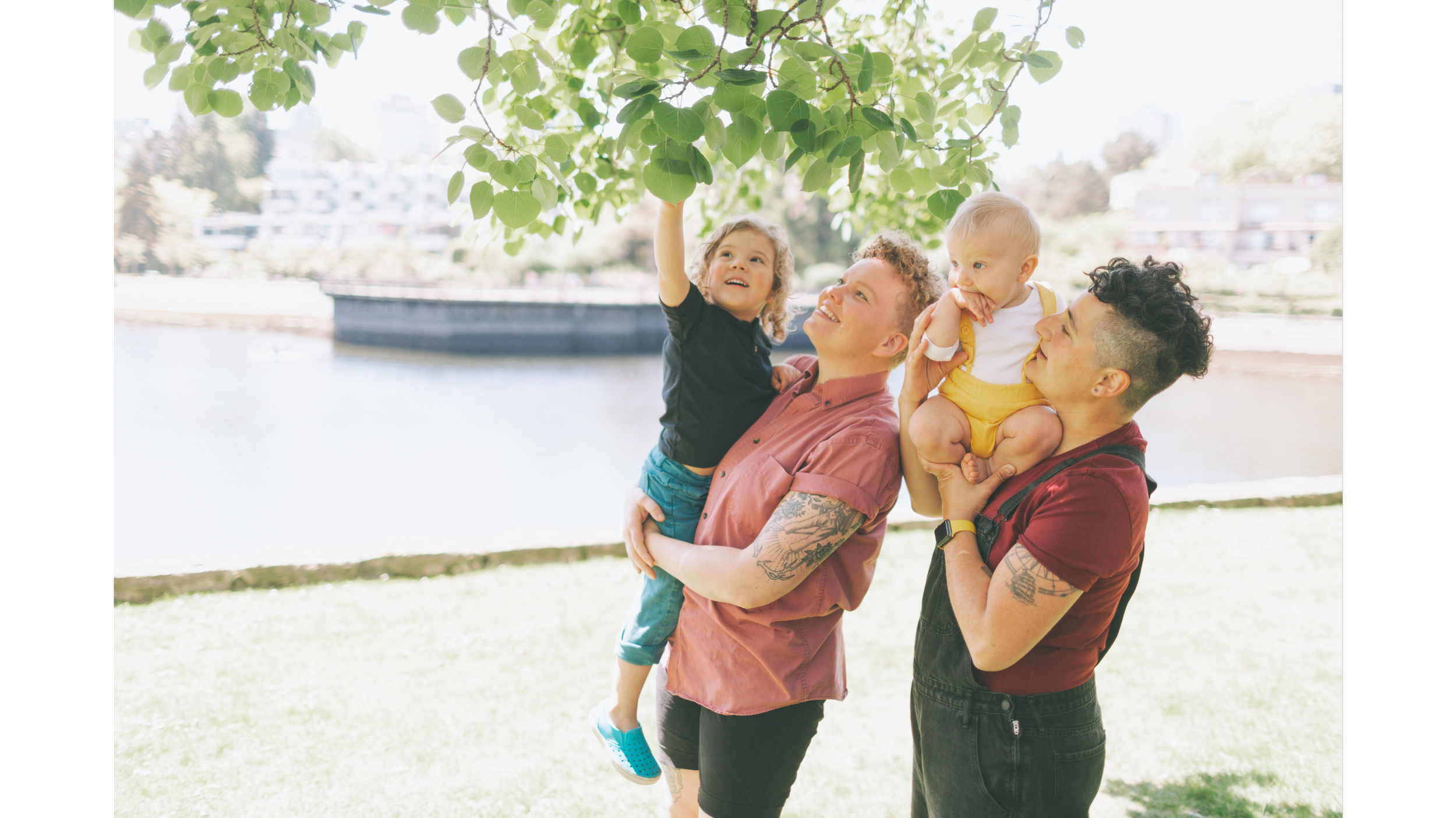 Two adults holding two children under a leafy tree next to a river, smiling and playing outside on a sunny day.