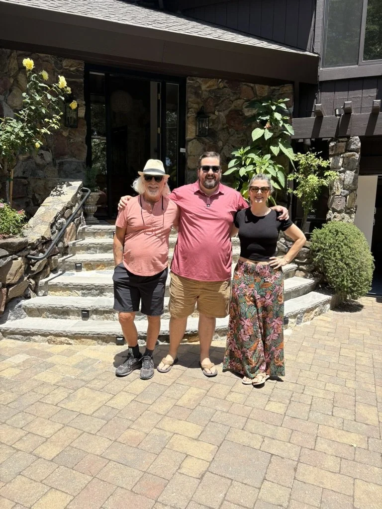 Three people standing outside a stone house, smiling and posing for a photo on a sunny day.
