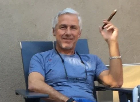An older man with gray hair and a beard, smiling while sitting in a chair, holding a cigar in his right hand, wearing a blue t-shirt.