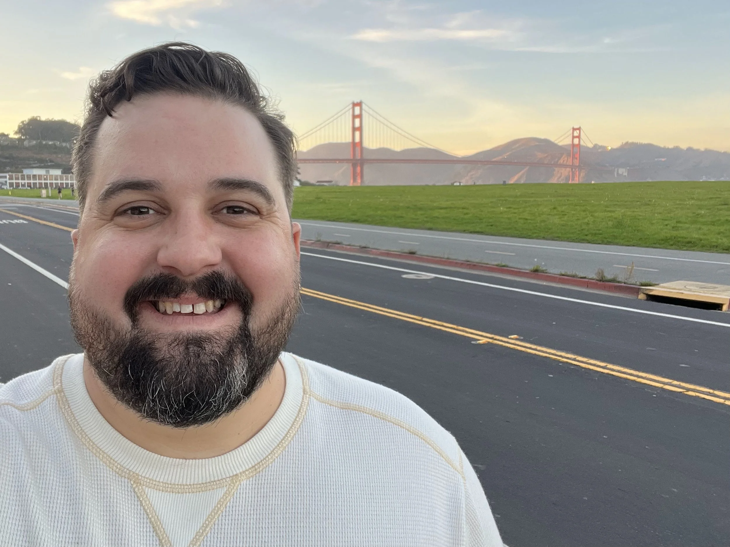 A man with dark hair, a beard, and a mustache smiling in front of the Golden Gate Bridge during sunset or sunrise with a partly cloudy sky.