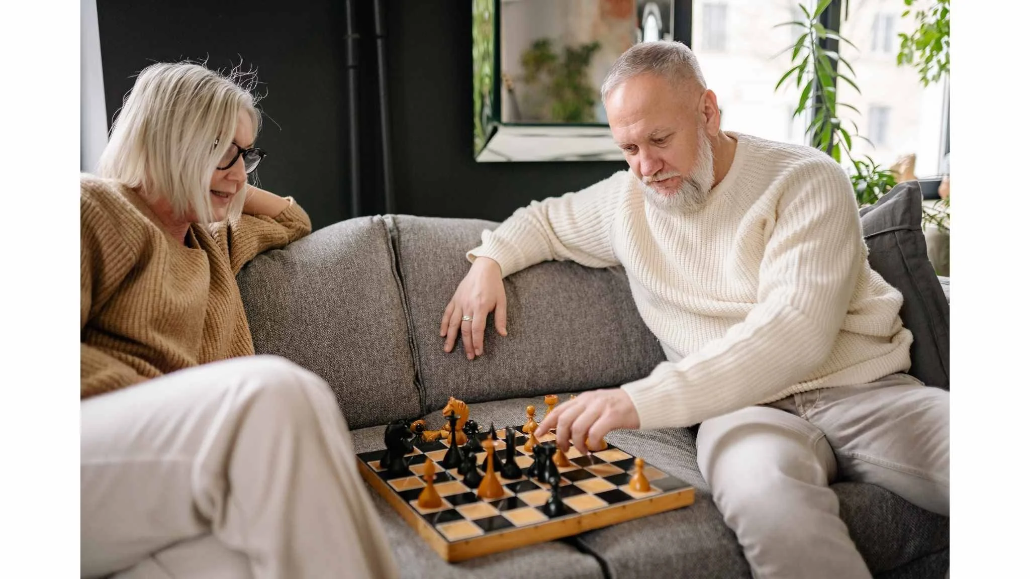 An elderly couple is playing chess on a gray couch in a cozy living room. The woman has shoulder-length blonde hair, wears glasses, and a brown sweater. The man has short gray hair, a beard, and is wearing a cream-colored sweater. They are focused on the game, with the man making a move and the woman watching.