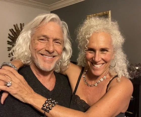 A happy older couple with white hair embracing each other and smiling at the camera indoors.