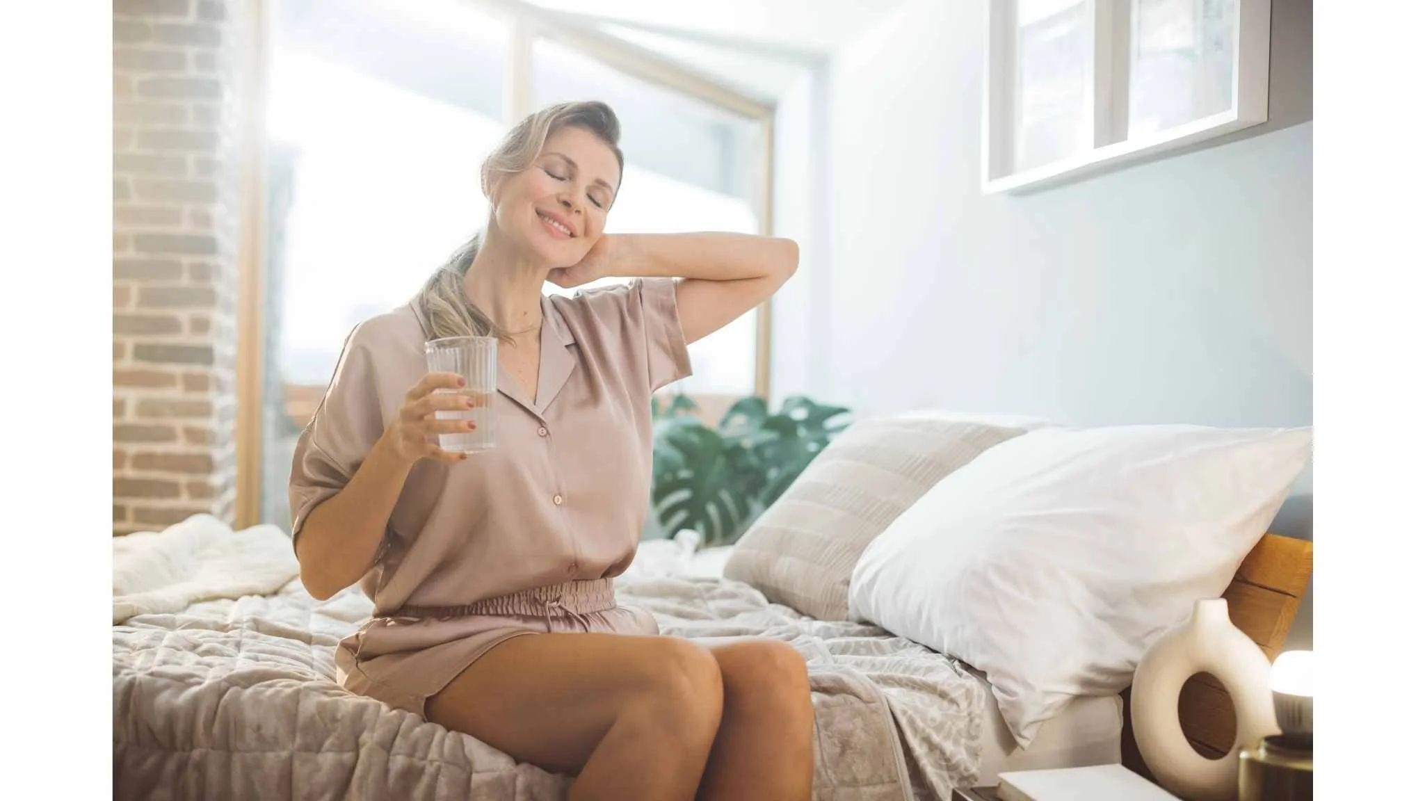 A woman sitting on a bed, smiling and holding a glass of water, in a bright and airy bedroom with a brick wall and large window.
