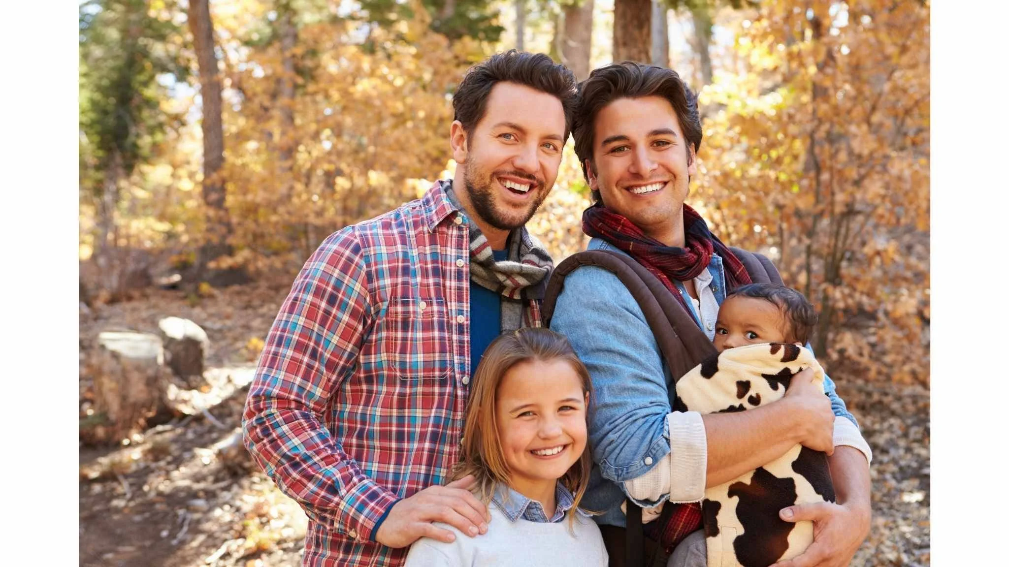 Two men and a young girl outdoors in a forest during autumn, smiling at the camera. One man is holding a baby wrapped in a cow-print blanket.