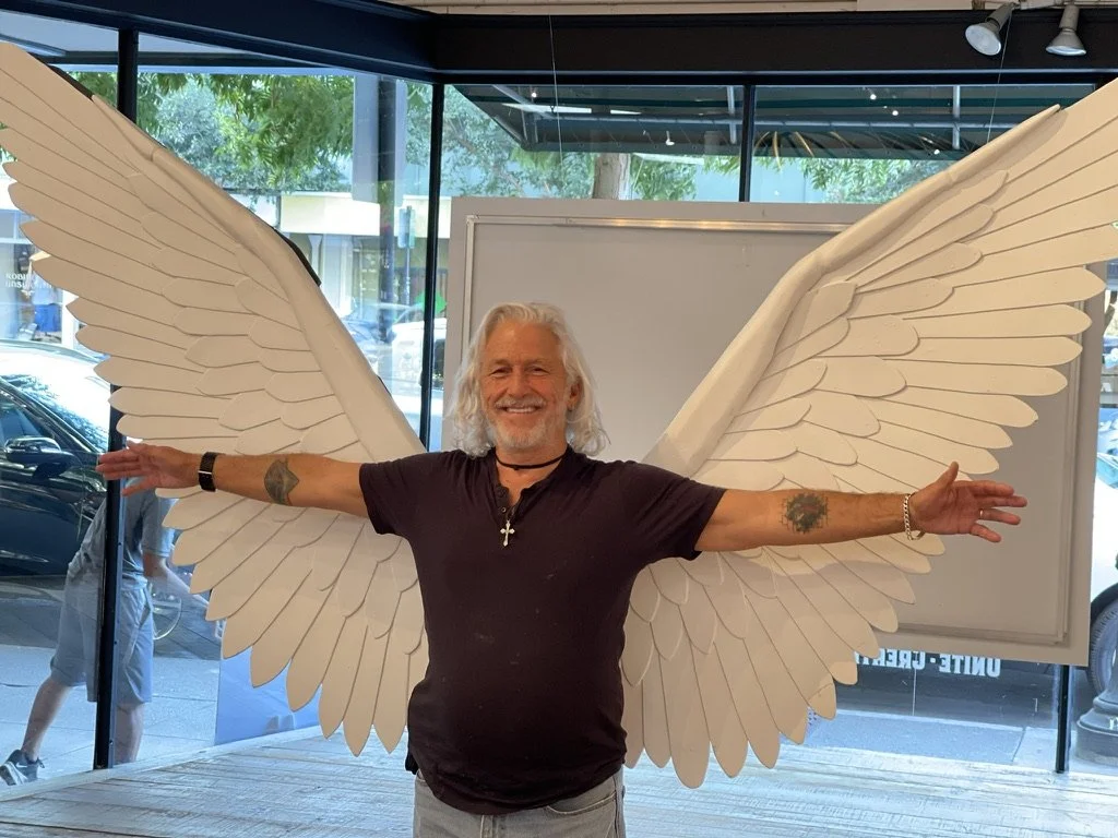 A man with white hair and a beard, smiling, stands in front of large white angel wings on a wall, with his arms outstretched.