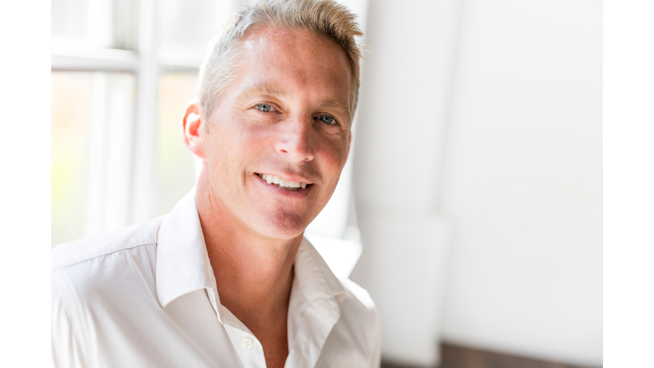 Close-up of smiling middle-aged man with blonde hair and blue eyes, wearing a white dress shirt, in a well-lit room with large windows in the background.