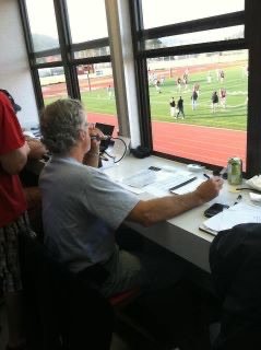Man sitting at a table inside a press box, looking out a window at a sports field with players and spectators.
