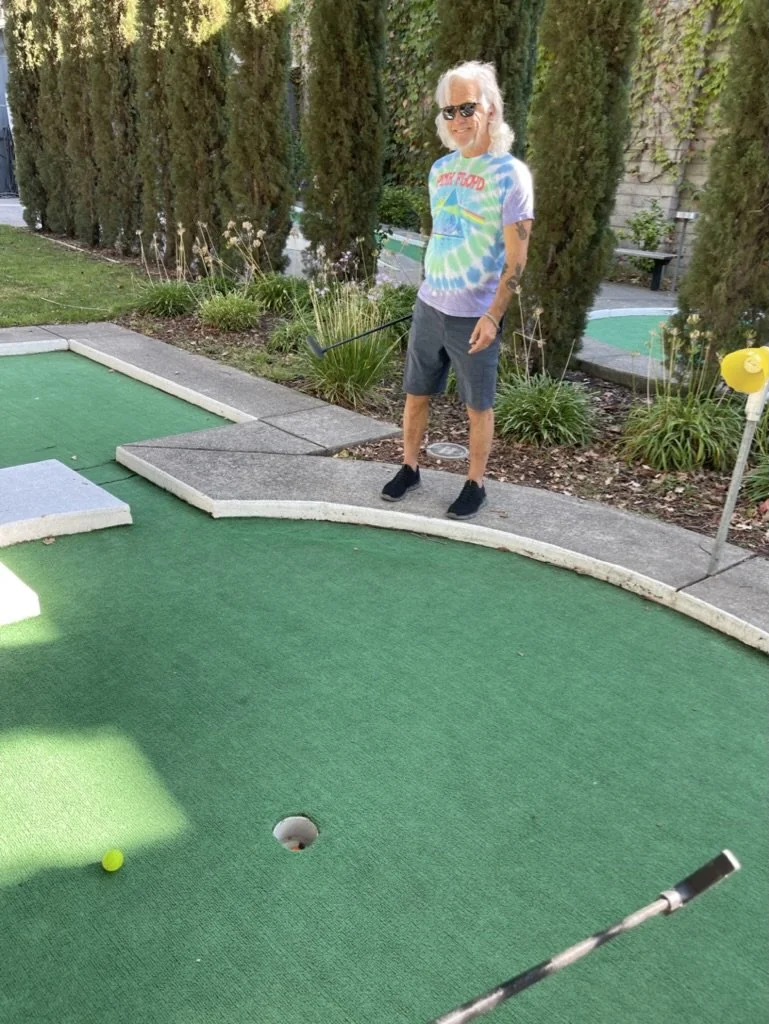 A woman with white hair, sunglasses, and a tie-dye shirt stands on a mini golf course, holding a putter, with a golf ball near the hole.