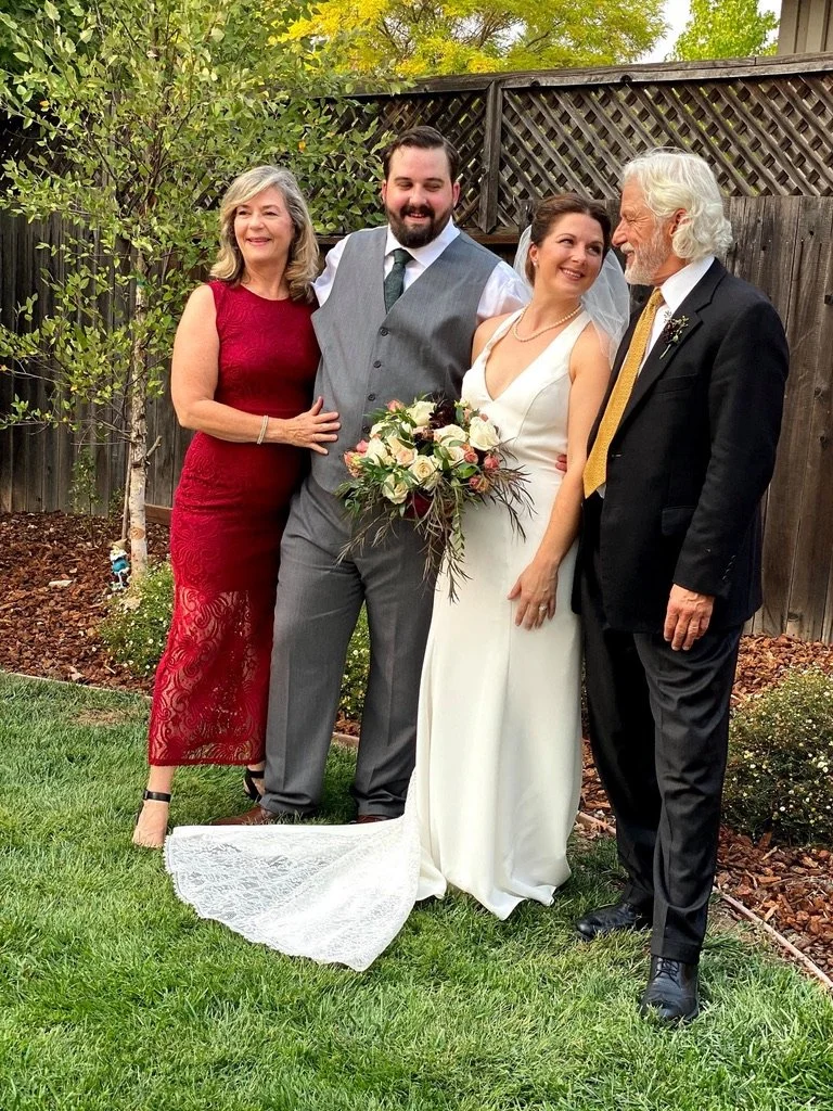 A group of four people, including a bride in a white wedding dress holding a bouquet, an older man in a black suit, a groom in a gray vest, and a woman in a red dress, standing outdoors in a garden with a wooden fence and trees in the background.