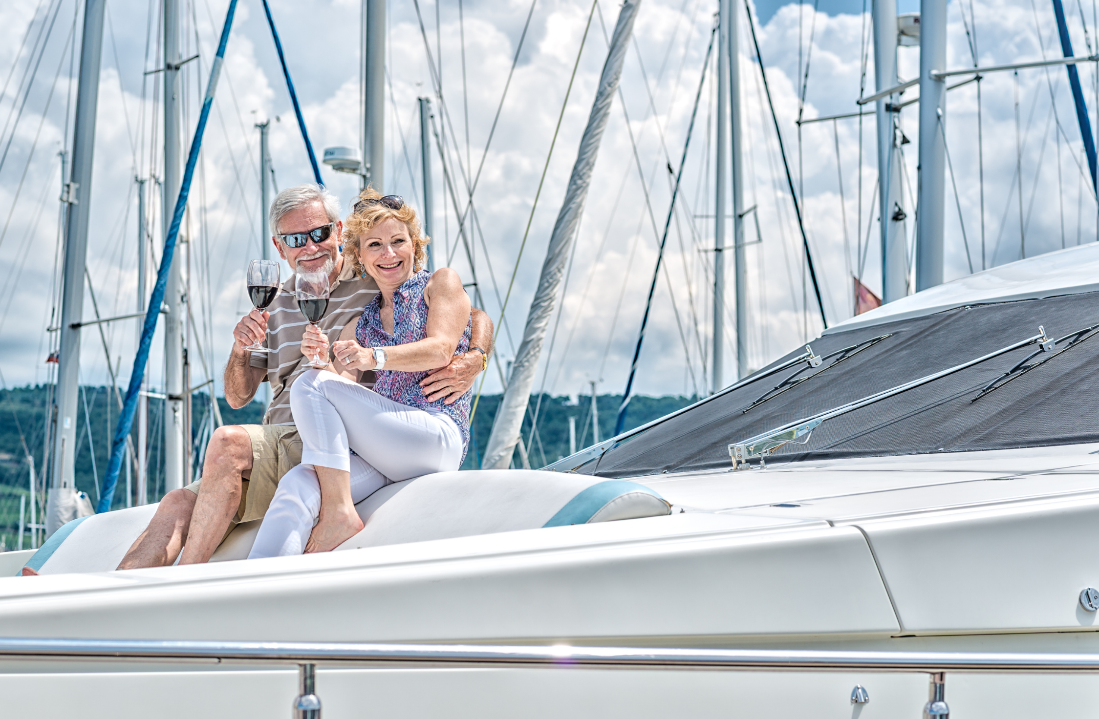 An elderly couple smiling and holding glasses of red wine while sitting on the deck of a white yacht at a marina with sailboats and cloudy sky in the background.