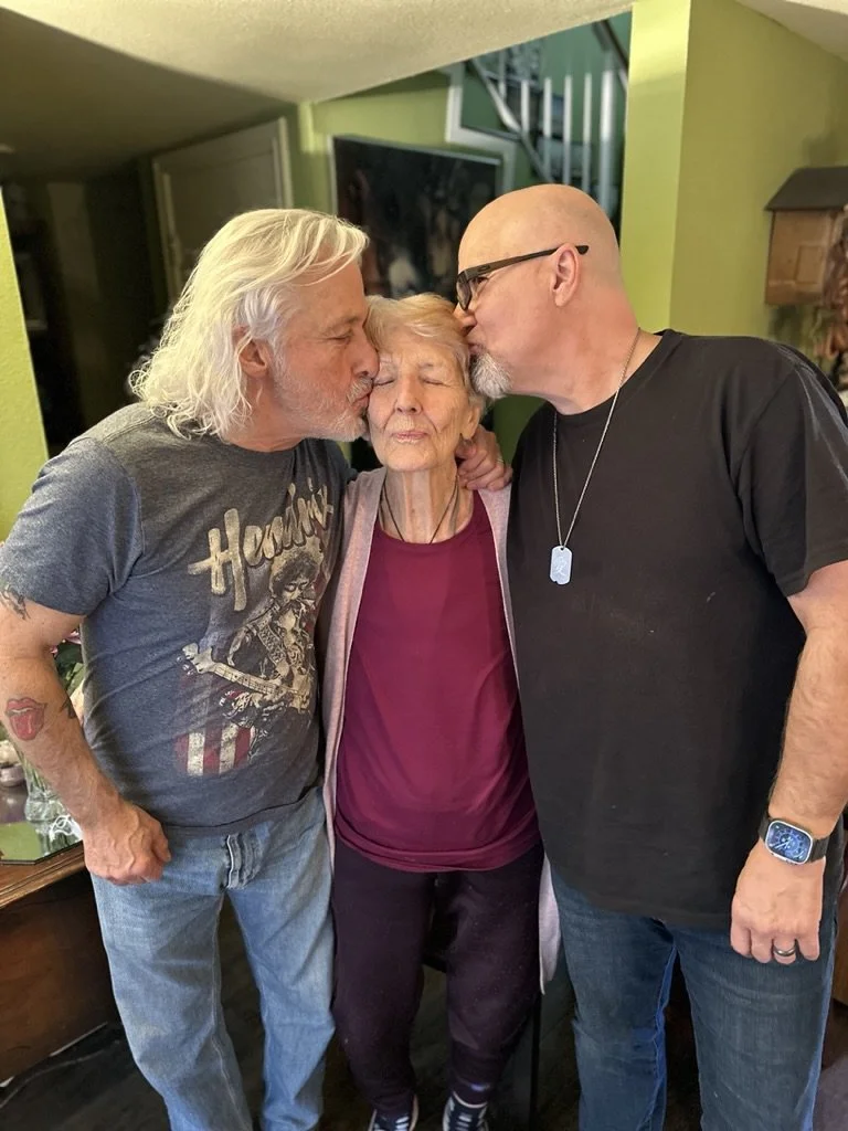 Two men kissing an elderly woman on the cheeks in a cozy, green-walled living room.