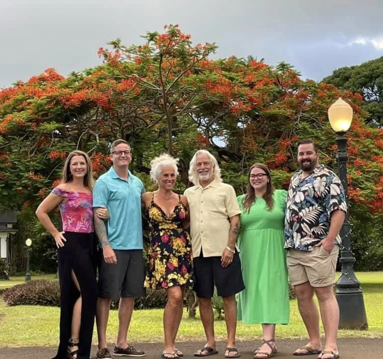 Group of six people standing outdoors in front of a large flowering tree, smiling, with a park setting and lamp post.