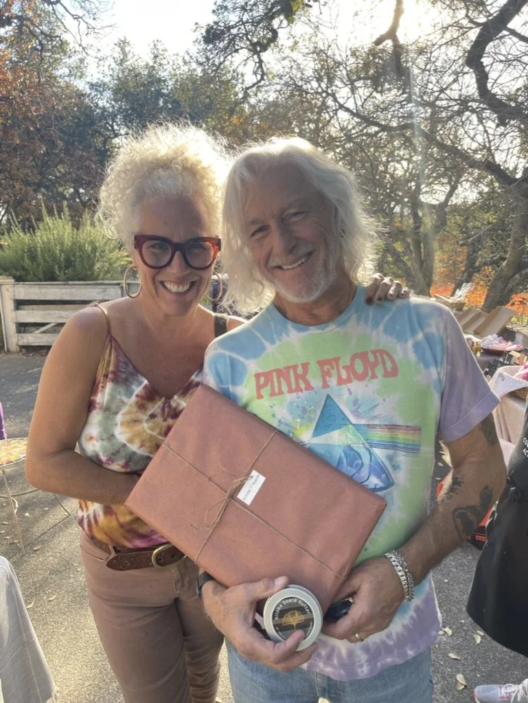 A woman with curly blonde hair, glasses, and a tie-dye tank top standing next to a man with long white hair, a Pink Floyd t-shirt, holding a wrapped gift and a small round object, outdoors on a sunny day.