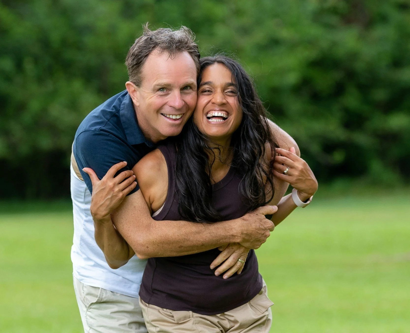 A happy middle-aged man with light skin and dark hair hugging a smiling middle-aged woman with dark hair and darker skin outdoors on a grassy area with trees in the background.